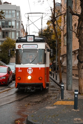A red and white tram is traveling on a city street. The tram has a sign displaying 'KADIKÖY - MODA' and the number '20'. A man is visible inside the tram, and there are buildings, cars, and trees lining the street. The street appears to be slightly wet, indicating recent rain.