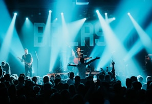 A live concert scene with a musician playing a guitar on stage, surrounded by bright lights and a cheering crowd. The background features large illuminated letters and the atmosphere is vibrant and energetic.