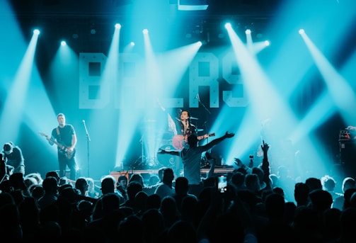 A live concert scene with a musician playing a guitar on stage, surrounded by bright lights and a cheering crowd. The background features large illuminated letters and the atmosphere is vibrant and energetic.