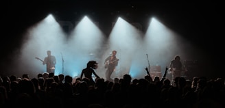 A concert scene with a band performing on stage under dramatic lighting. The silhouettes of four musicians are visible, each holding a guitar. A crowd is gathered in front of the stage, with some people raising their hands in excitement.