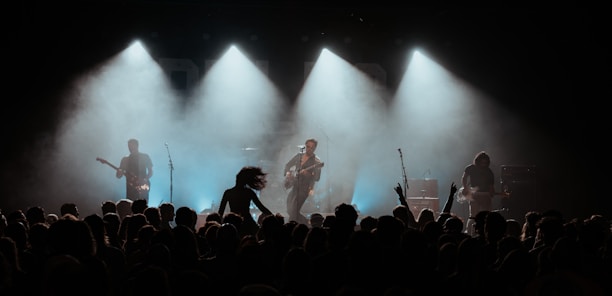 A concert scene with a band performing on stage under dramatic lighting. The silhouettes of four musicians are visible, each holding a guitar. A crowd is gathered in front of the stage, with some people raising their hands in excitement.