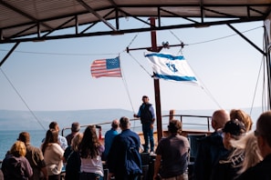 A group of people stands on a boat under a metal roof, facing a man who appears to be speaking. The American flag and Israeli flag are prominently displayed, waving in the breeze. The background reveals a body of water with foggy mountains beyond.