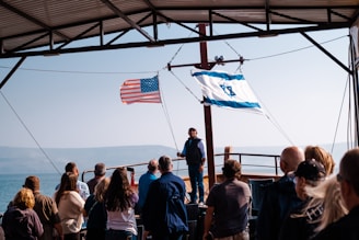 A group of people stands on a boat under a metal roof, facing a man who appears to be speaking. The American flag and Israeli flag are prominently displayed, waving in the breeze. The background reveals a body of water with foggy mountains beyond.