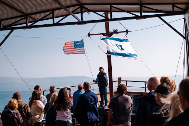 A group of people stands on a boat under a metal roof, facing a man who appears to be speaking. The American flag and Israeli flag are prominently displayed, waving in the breeze. The background reveals a body of water with foggy mountains beyond.