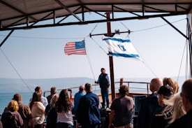 A group of people stands on a boat under a metal roof, facing a man who appears to be speaking. The American flag and Israeli flag are prominently displayed, waving in the breeze. The background reveals a body of water with foggy mountains beyond.