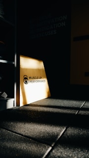 A dimly lit gym environment with a focus on a wooden panel displaying 'Muscle Up Performance'. Shadows and light create a dramatic effect on the dark-tiled floor. Motivational words like 'Focus', 'Motivation', 'Dedication', and 'Excuses' are subtly visible on the wall.
