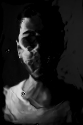 A distorted and abstract representation of a person emerges from a dark, fluid-like background. The image features a mix of light and shadow, creating an obscured, ghostly appearance with a soft focus on certain facial features. The interplay of light highlights parts of the face, adding a mysterious and surreal quality.