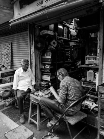 Two older men are engaged in conversation outside a small, cluttered shop filled with electronic equipment. One man sits on a stool while the other occupies a folding chair. The shop is packed with various items including radios and other electronic devices.