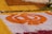 An intricate floral pattern is arranged on the ground using a variety of colorful petals. A prominent orange design is set against a white background, surrounded by sections of yellow and red petals. Green leaves add contrast and texture. There are also clusters of white flowers and some visible greenery.