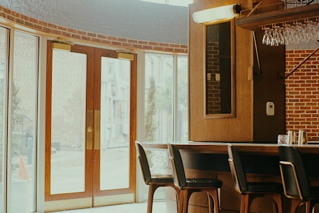 A cozy interior of a bar or cafe featuring a wooden bar counter with several high chairs. Glasses are hanging upside down above the counter. Large glass doors with wooden frames allow natural light to flood the space, and the walls are adorned with a red brick design.