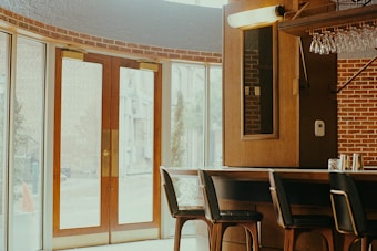 A cozy interior of a bar or cafe featuring a wooden bar counter with several high chairs. Glasses are hanging upside down above the counter. Large glass doors with wooden frames allow natural light to flood the space, and the walls are adorned with a red brick design.