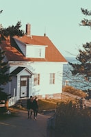A cozy house with a red roof and white wooden siding is situated near the coastline. Two people are walking on the path leading up to the house. The setting is surrounded by trees, with a view of the sea in the background.