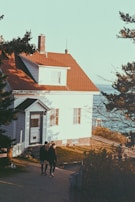 A cozy house with a red roof and white wooden siding is situated near the coastline. Two people are walking on the path leading up to the house. The setting is surrounded by trees, with a view of the sea in the background.