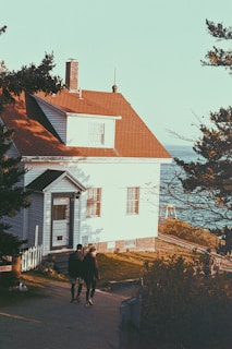 A cozy house with a red roof and white wooden siding is situated near the coastline. Two people are walking on the path leading up to the house. The setting is surrounded by trees, with a view of the sea in the background.