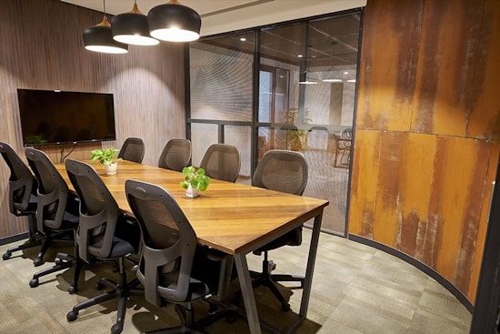 A modern conference room with a long wooden table surrounded by eight black mesh office chairs. Three black pendant lights hang above the table, illuminating the room. A flat-screen TV is mounted on a wooden paneled wall, and there are small green potted plants placed on the table. The room has glass partition walls and rust-colored decorative panels.