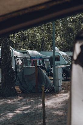A row of caravans is parked in a wooded campsite. There is a tree trunk in the foreground and a road sign with the text 'Australijska' visible. A car and several covered items are nearby, with a power box and some electrical cables also present.