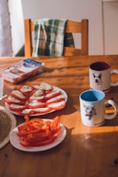 A wooden table features two ceramic mugs each adorned with animal illustrations, one with a blue inside and another with a red inside. A green and yellow checkered kitchen towel is draped over the back of a wooden chair. Plates on the table hold slices of tomato topped with mozzarella and herbs, and sliced red bell peppers. A package of what appears to be sliced meat is next to the plates.