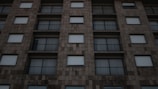 An urban building facade with a grid-like pattern of square windows and metal railings. The building's stone-like exterior appears uniform and structured, featuring muted, earthy tones.