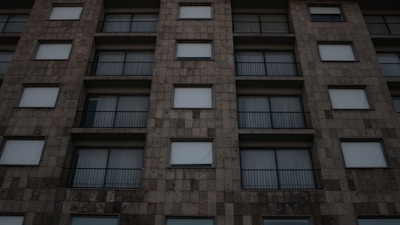An urban building facade with a grid-like pattern of square windows and metal railings. The building's stone-like exterior appears uniform and structured, featuring muted, earthy tones.