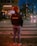 A person with curly hair stands on a crosswalk at night, wearing a jacket with 'Café do Brazil' written on the back. The street is illuminated by streetlights and headlights from cars, with some pedestrians in the background near a building with a sign reading 'Kitanda'.