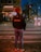 A person with curly hair stands on a crosswalk at night, wearing a jacket with 'Café do Brazil' written on the back. The street is illuminated by streetlights and headlights from cars, with some pedestrians in the background near a building with a sign reading 'Kitanda'.
