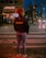 A person with curly hair stands on a crosswalk at night, wearing a jacket with 'Café do Brazil' written on the back. The street is illuminated by streetlights and headlights from cars, with some pedestrians in the background near a building with a sign reading 'Kitanda'.