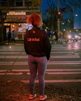 A person with curly hair stands on a crosswalk at night, wearing a jacket with 'Café do Brazil' written on the back. The street is illuminated by streetlights and headlights from cars, with some pedestrians in the background near a building with a sign reading 'Kitanda'.