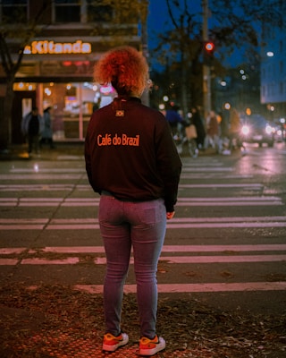 A person with curly hair stands on a crosswalk at night, wearing a jacket with 'Café do Brazil' written on the back. The street is illuminated by streetlights and headlights from cars, with some pedestrians in the background near a building with a sign reading 'Kitanda'.