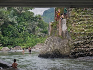 A group of people are gathered on a large rock near a river, with lush green tropical vegetation and trees in the background. Some individuals are in the water, while others are sitting or standing on the rock structure, which has a rough, textured surface. The scene suggests a recreational or leisure activity taking place in a natural setting.