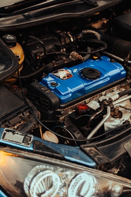A close-up view of a car engine compartment displaying a blue engine cover with surrounding mechanical components. Various hoses and wires are visible, along with the headlight structure at the foreground.