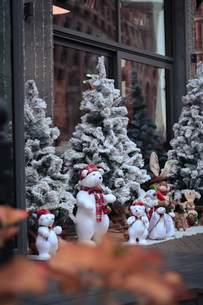 A festive display featuring snow-covered Christmas trees and plush figures of polar bears dressed in red and black checkered scarves and hats. The scene is set in front of a window, likely a storefront, reflecting some urban architecture. Additional plush toys, including rabbits and other small animals, are also part of the display.