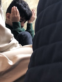 A young child sitting among adults, with hands raised to the sides of their face, creating a sense of contemplation or concentration. The child appears to be in a thoughtful or meditative pose.