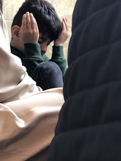 A young child sitting among adults, with hands raised to the sides of their face, creating a sense of contemplation or concentration. The child appears to be in a thoughtful or meditative pose.