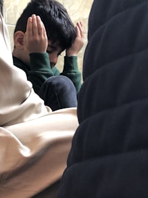 A young child sitting among adults, with hands raised to the sides of their face, creating a sense of contemplation or concentration. The child appears to be in a thoughtful or meditative pose.