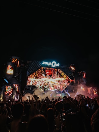 A vibrant concert scene with a large crowd raising their hands in front of a brightly lit stage. The stage features colorful LED screens displaying dynamic visuals with orange and red hues. The atmosphere is energized, and a thick haze of smoke adds to the dramatic lighting effects.
