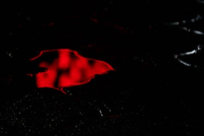 A dark setting with a glossy surface reflecting a vibrant red pool of liquid. The intense color contrasts sharply with the surrounding darkness, creating a mysterious and ominous atmosphere.
