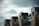 A series of brick chimneys with dark caps are aligned on the roof of a building. Several seagulls are perched atop the chimneys, and the sky is overcast with a mix of clouds, creating a dramatic backdrop.