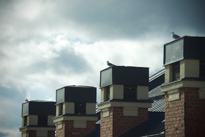 A series of brick chimneys with dark caps are aligned on the roof of a building. Several seagulls are perched atop the chimneys, and the sky is overcast with a mix of clouds, creating a dramatic backdrop.