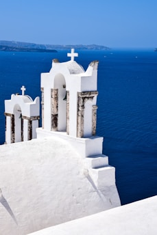 A white church with a cross on top, set against a vivid blue seascape. The architecture is traditional, with smooth white surfaces and arched structures. The background includes the vast blue sea and a distant coastline under a clear sky.