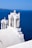 A white church with a cross on top, set against a vivid blue seascape. The architecture is traditional, with smooth white surfaces and arched structures. The background includes the vast blue sea and a distant coastline under a clear sky.