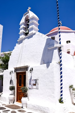 A traditional whitewashed building with a wooden door and two potted plants on either side. The structure features a bell and a cross on top, characteristic of Greek island architecture. A striped flagpole stands beside it, with notices posted near the entrance. The bright blue sky provides a vivid contrast to the white facade.