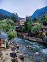 A serene mountain landscape features a flowing river with clear blue waters surrounded by rocks and lush green vegetation. On the riverbank, there are colorful buildings, including a multi-story structure with a pink roof. The scene is enveloped by forested hills and towering mountains in the background.
