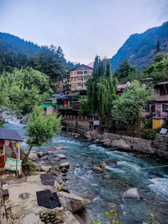 A serene mountain landscape features a flowing river with clear blue waters surrounded by rocks and lush green vegetation. On the riverbank, there are colorful buildings, including a multi-story structure with a pink roof. The scene is enveloped by forested hills and towering mountains in the background.
