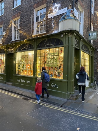 A cozy street scene featuring a shop with green-painted exterior and large windows, displaying various colorful items and Christmas decorations. The warm glow of fairy lights and a large star adorn the upper part of the building. Two people walk by on the pavement, one wearing a red coat and the other a backpack. A sign reads 'High Petergate' and another, 'Fair Trade, Eco, Global Gifts'.