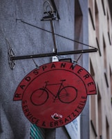 A circular red sign with a black illustration of a bicycle is affixed to a wall. The sign has text in German, 'Radspannerei Service & Ersatzteile,' indicating a bicycle service and parts shop. The metal frame holding the sign has a gear-like element on the side.