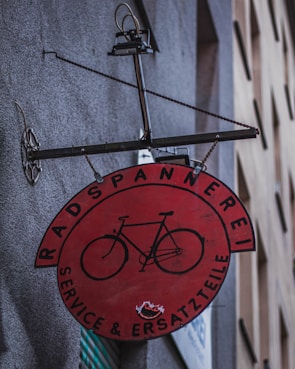 A circular red sign with a black illustration of a bicycle is affixed to a wall. The sign has text in German, 'Radspannerei Service & Ersatzteile,' indicating a bicycle service and parts shop. The metal frame holding the sign has a gear-like element on the side.