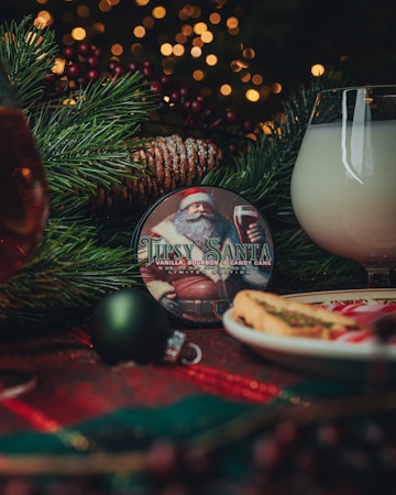 A cozy holiday scene featuring a decorative round object with an illustration of Santa Claus and the text 'Tipsy Santa' on it. Surrounding elements include a pine branch with a dusting of snow, Christmas ornaments, a glass of milky beverage, and a plate with cookies. Warm, blurred lights in the background add to the festive atmosphere.
