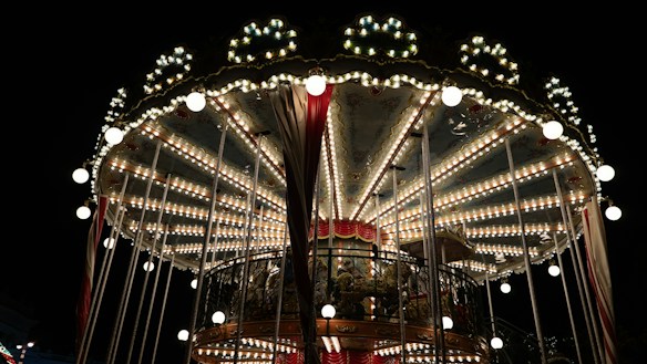 A brightly lit carousel against a dark night sky, featuring intricate decorative details and illuminated light bulbs. The design includes ornate patterns and a vibrant, festive appearance with red and gold accents.