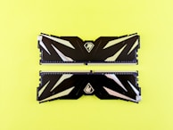 Two black RAM sticks featuring geometric patterns and metallic accents are placed side by side on a bright yellow background. Each RAM stick displays a sleek design with a logo in the center.