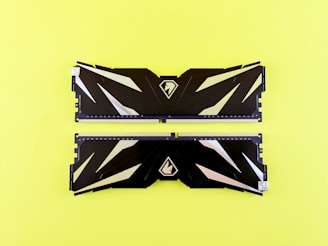 Two black RAM sticks featuring geometric patterns and metallic accents are placed side by side on a bright yellow background. Each RAM stick displays a sleek design with a logo in the center.
